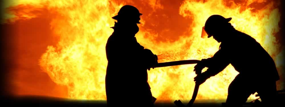 Silhouettes of two people in helmets working with a hose against a massive orange fire, dramatic high-contrast mood.