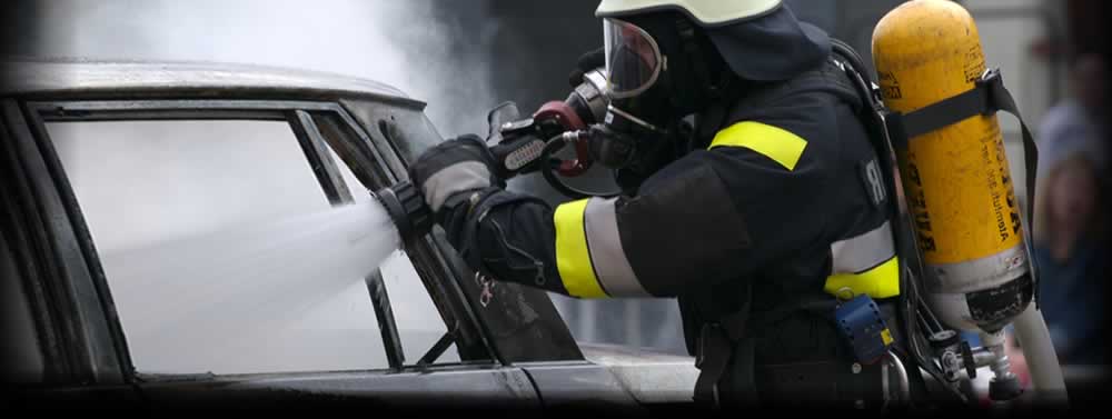 Person in protective gear and breathing apparatus spraying water into a smoking vehicle window, muted colors, tense action scene.